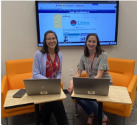 Two people sit on orange chairs with their laptops open. They are smiling at the camera.
