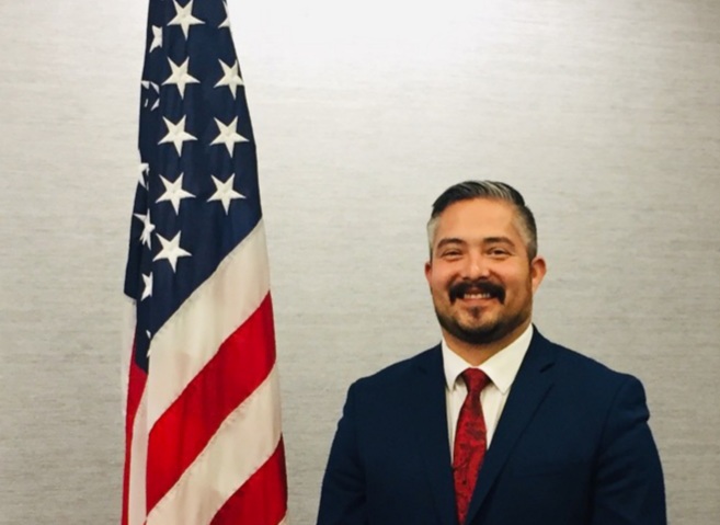 Nick Lopez Man with dark hair, mustache, and beard in a blue suit with a red tie standing next to an american flag