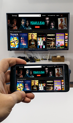 A hand holds a phone with a mall-mounted TV in the background. Both screens share the same content which is a display of video titles for checkout.