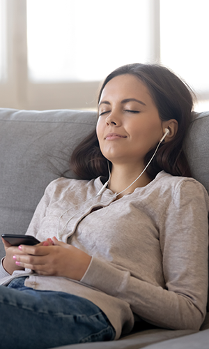 A woman relaxing on the couch while she listens to an audiobook playing from a mobile phone in her hand.