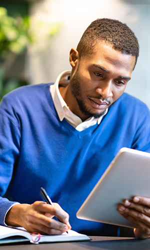 A man in a blue sweater takes notes ina notebook while holding a tablet in the other hand.