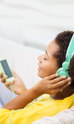 A woman in a yellow shirt, eyes closed and smiling, has a hand placed up to one of the headest speakers shew ears that is the same matching mint color as the phone her headset connects to via wire.