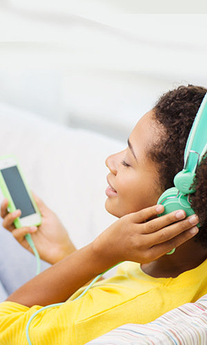 A woman in a yellow shirt, eyes closed and smiling, has a hand placed up to one of the headest speakers shew ears that is the same matching mint color as the ohone her headset connects to via wire.