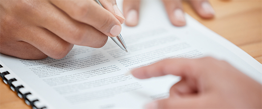 A Hand with a pen hovering above a document with the index finger of another individual pointing at the document