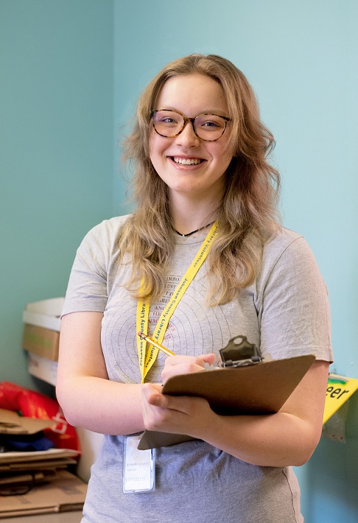 A person holding a clipboard and wearing a yellow lanyard smiles at the camera