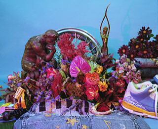 Still life arrangement of tropical and dried plants, bronze statues, hubcap, denim and a sneaker.