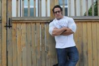 A person wearing sunglasses and a white shirt and jeans leans with their arms crossed against a picket fence.