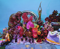 Photograph of still life arrangement of tropical and dried plants, bronze statues, a hubcap, denim and sneaker.