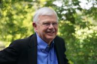 A smiling white haired person in a blue shirt and dress jacket stands in front of trees