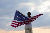 Woman stands with her back to the camera, holding an american flag behind her and draped across her back
