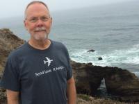 Dennis Ross posing in front of a large body of water with a rocky shore