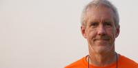 John Siceloff poses in an orange shirt in front of a plan light background