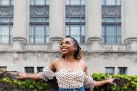 A black woman with her arms open wide leaning against an ivy covered wall looking off to the side and smiling wide.