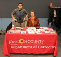 a smiling standing person and a smiling seated person behind a table with a red 'department of corrections' cloth and many flyers and handouts on top