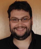 A person in a black shirt and glasses, with a beard, smiling into the camera.