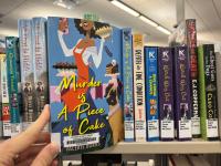 A hand pulling the book Murder is a Piece of Cake off of a shelf of other books