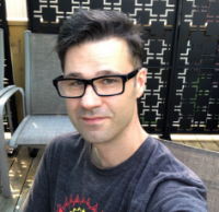 a person with short dark hair and rectangular glasses, wearing a gray t-shirt, takes a selfie