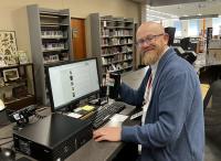 matt imrie poses at the front desk of the gardner library, with the computer next to him and the rest of the library in the background