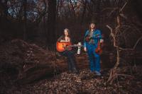 Holly Taylor and Buck Brandt of Folk in the Flow stand with their instruments, surrounded by trees.