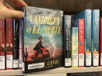 The book Vampires of El Norte being pulled off of a bookshelf of other books with Library spine labels
