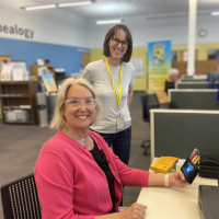 A person with shoulder length blond hair and a pink cardigan sits at a computer with their cell phone. Another person with shoulder length brown hair and a yellow volunteer lanyard stands near them, smiling at the camera