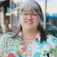 A person with shoulder length silver hair and bangs smiles at the camera. They are wearing glasses and a colorful button down shirt.
