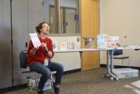 A person in jeans and a red sweater holds a book open to their audience, appearing to be reading the book out loud