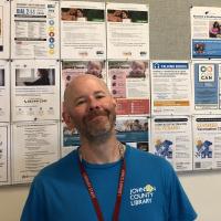 A person in a blue shirt and Johnson County Library lanyard stands in front of a community information board with flyers on it