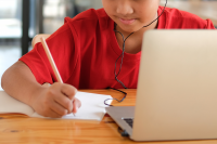 Photo of a boy in a red shirt writing notes with a pencil while listening to content from the open laptop in front of them.