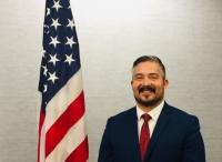 Man with dark hair, mustache, and beard in a blue suit with a red tie standing next to an american flag