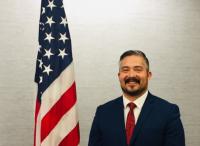 Nick Lopez Man with dark hair, mustache, and beard in a blue suit with a red tie standing next to an american flag