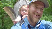 blonde bearded man in ball cap and blue shirt with baby in a backpack holding a feather