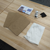 spider take and make kit Table with wooden top. On the table, printed instructions, foam paintbrush and black paint bottle, cardboard parts sheets, cotton webbing.