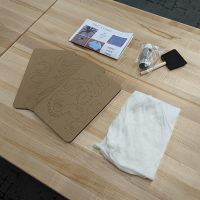 Table with wooden top. On the table, printed instructions, foam paintbrush and black paint bottle, cardboard parts sheets, cotton webbing.