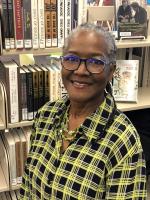 smiling black woman with glasses and a yellow and black checkered shirt in front of a book case