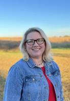 blond woman with shoulder length hair and glasses in a jean jacket with red shirt with field and sky behind