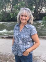 smiling woman with shoulder length grey hair in a blue short sleeved top with one hand in her jeans pocket