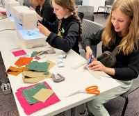 Two girls working at a sewing machine. One is running the machine while the other cuts fabric.