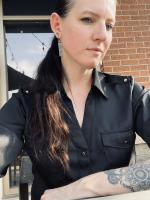 Morgan Schneebeli white woman with long dark hair in an over the shoulder ponytail in a black button up shirt with a collar and tatoos on her forarm