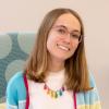 Natalie Wolf Young smiling woman with glasses, straight blondish hair, wearing a white t-shirt, sweater, and colorful necklace
