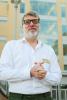 Man in white button down shirt with clasped hands, smiling, black framed glasses and a white beard